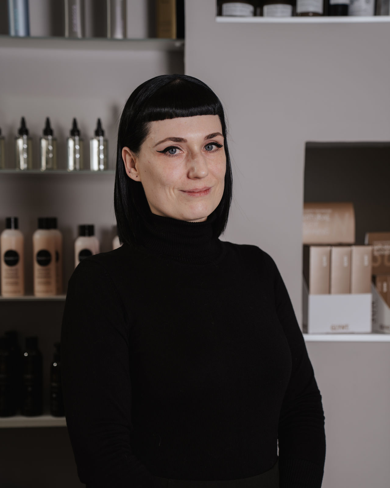 Mandy Startseite Friseurin Mandy steht im Salon Helena Conzen Hairdesign in Bochum vor einem Regal mit Haarpflegeprodukten.