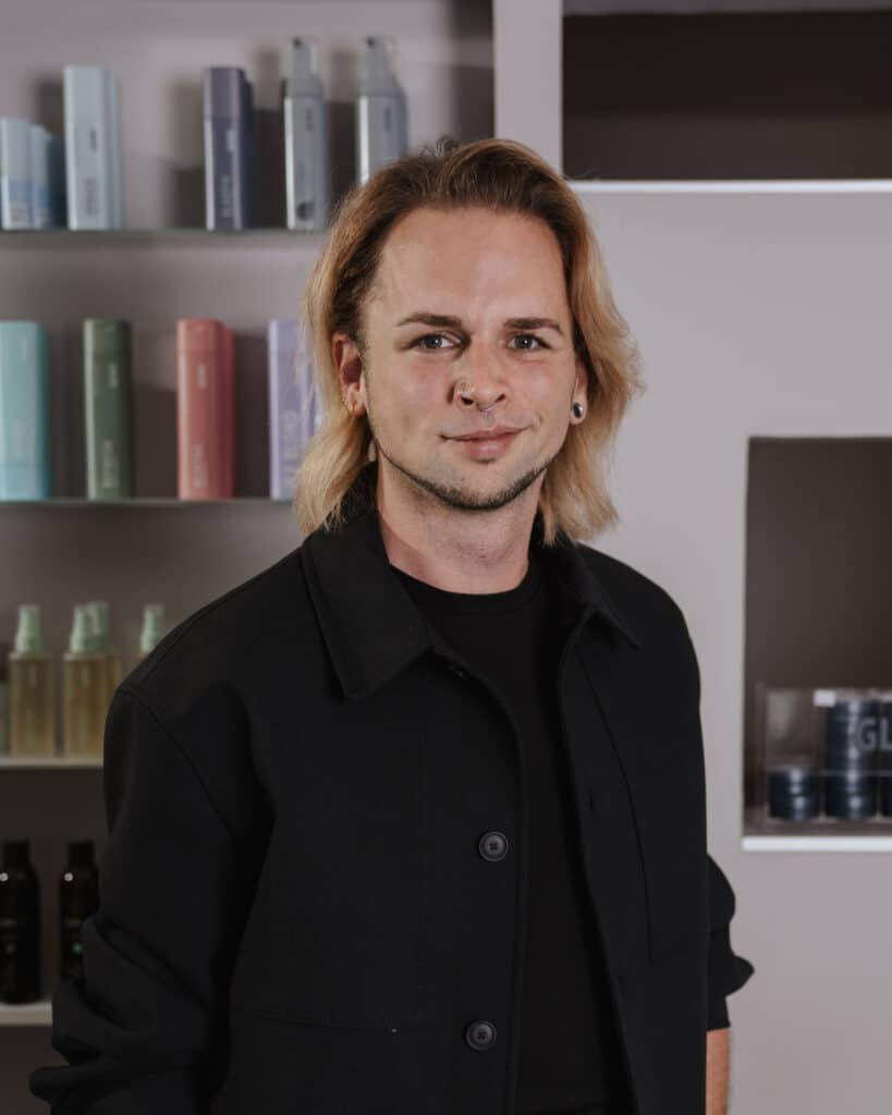 Daniel Willkommen im Team: Friseurmeister Daniel startet am 01.11. Friseur Daniel im Salon von Helena Conzen Hairdesign in Bochum vor einem Regal mit Haarpflegeprodukten